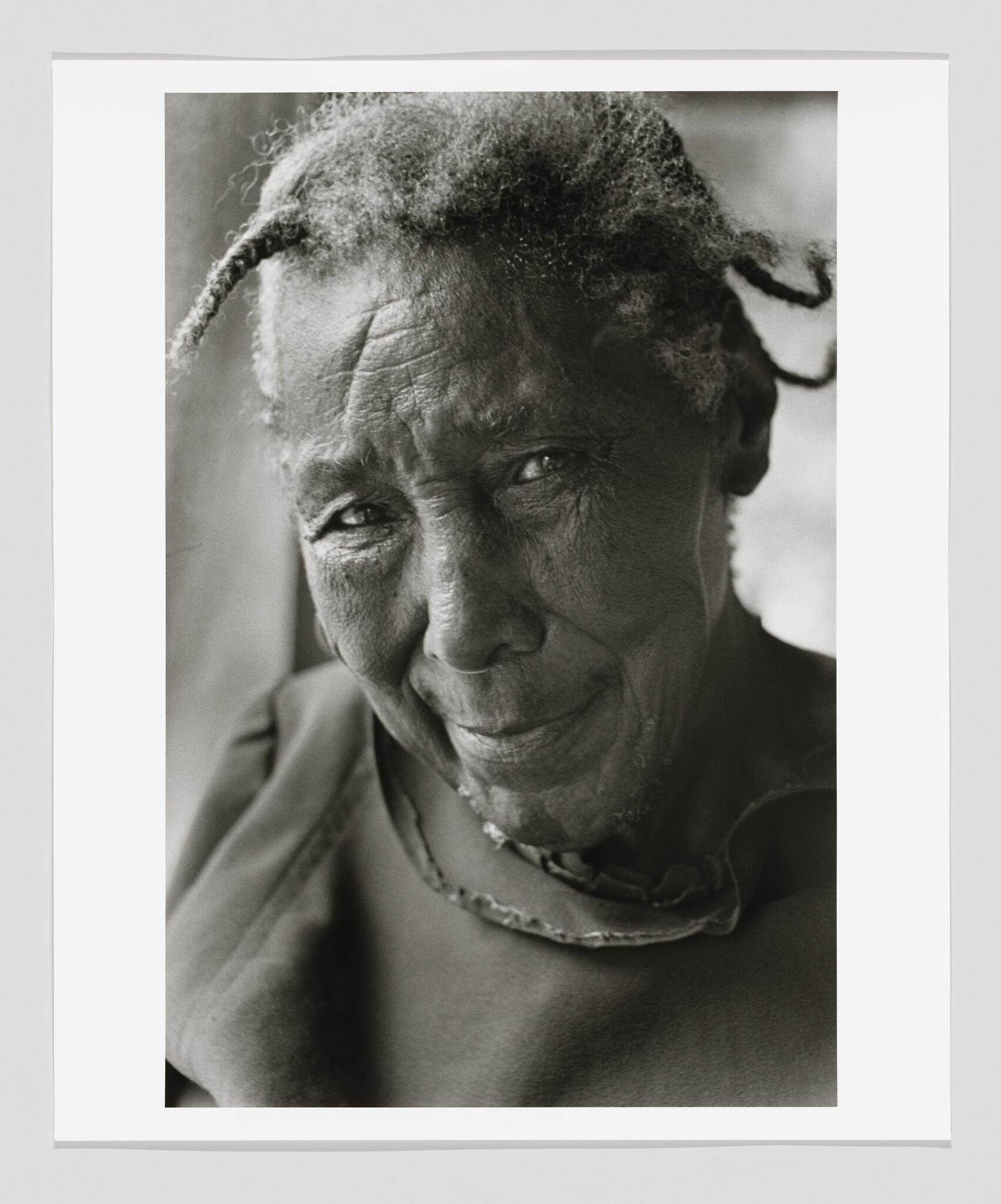 Elderly woman with textured hair looks toward the camera with a gentle, knowing smile.