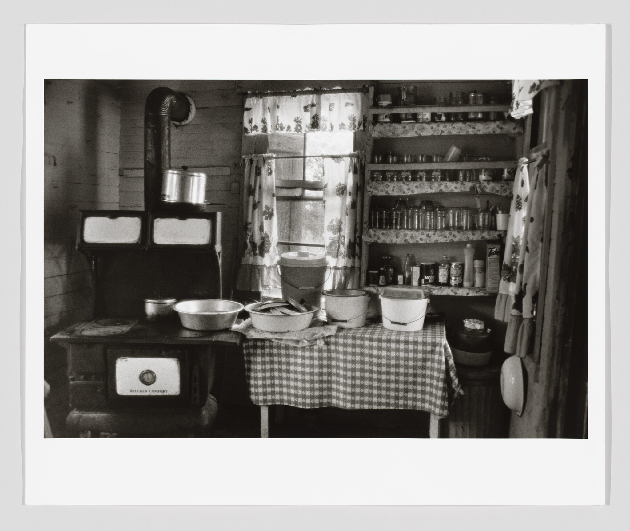An old country kitchen with a wood-burning stove, pots on a table, and shelves of jars.