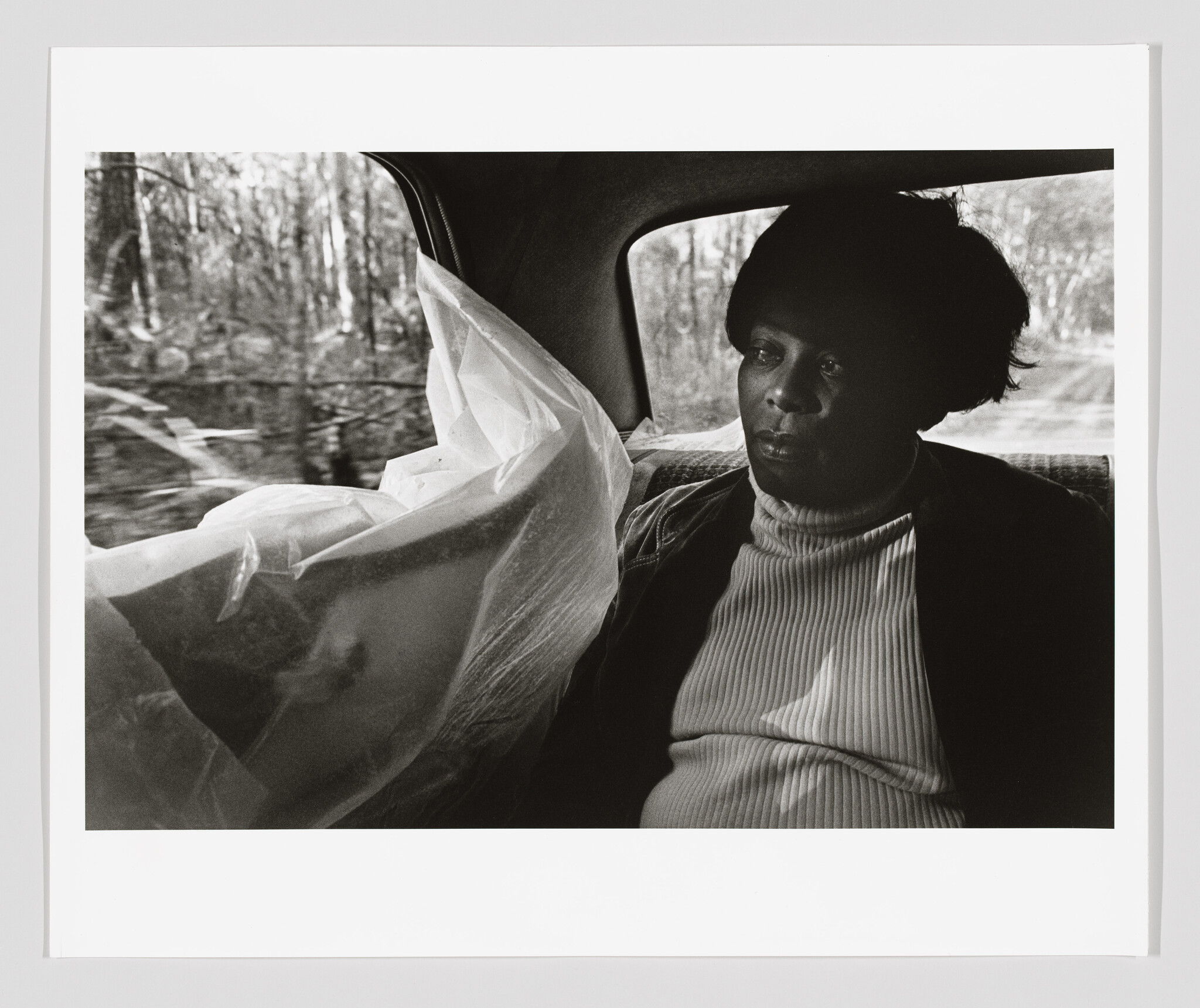 A woman sits in the backseat of a car, looking pensively out the window beside a plastic sheet.