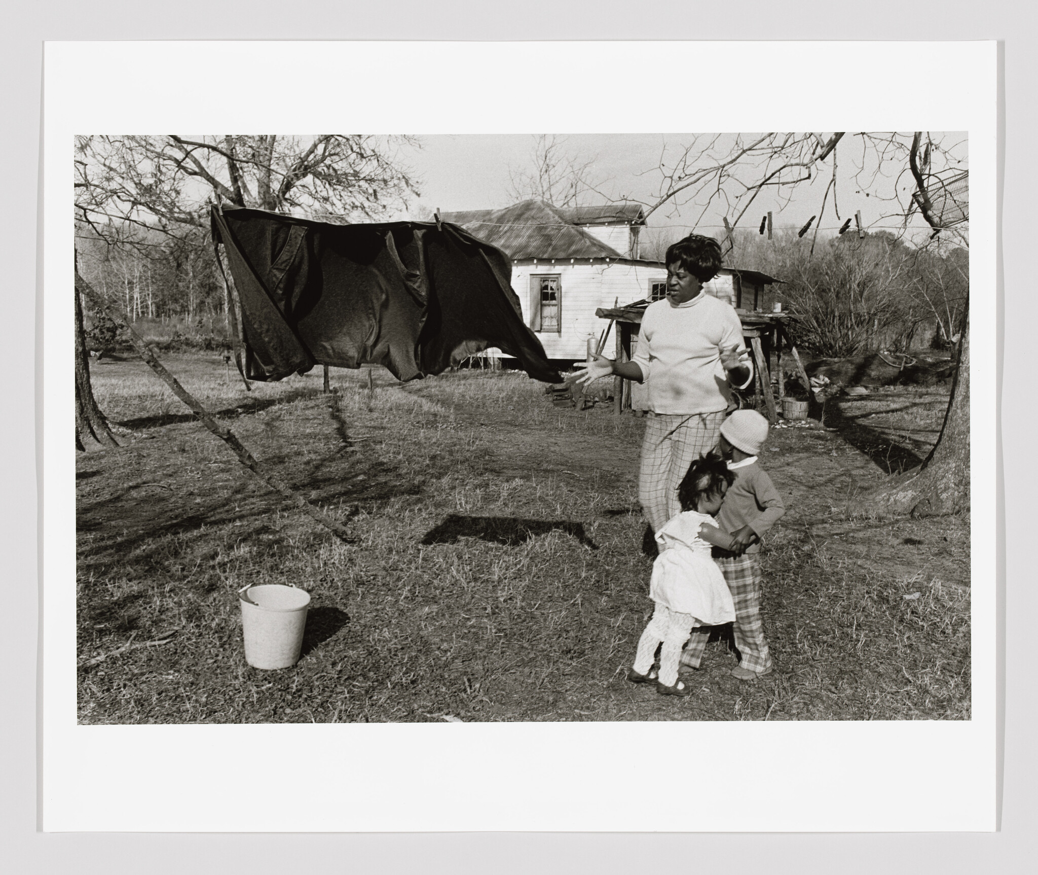 Woman hangs a large sheet on an outdoor clothesline while two children cling to her legs.