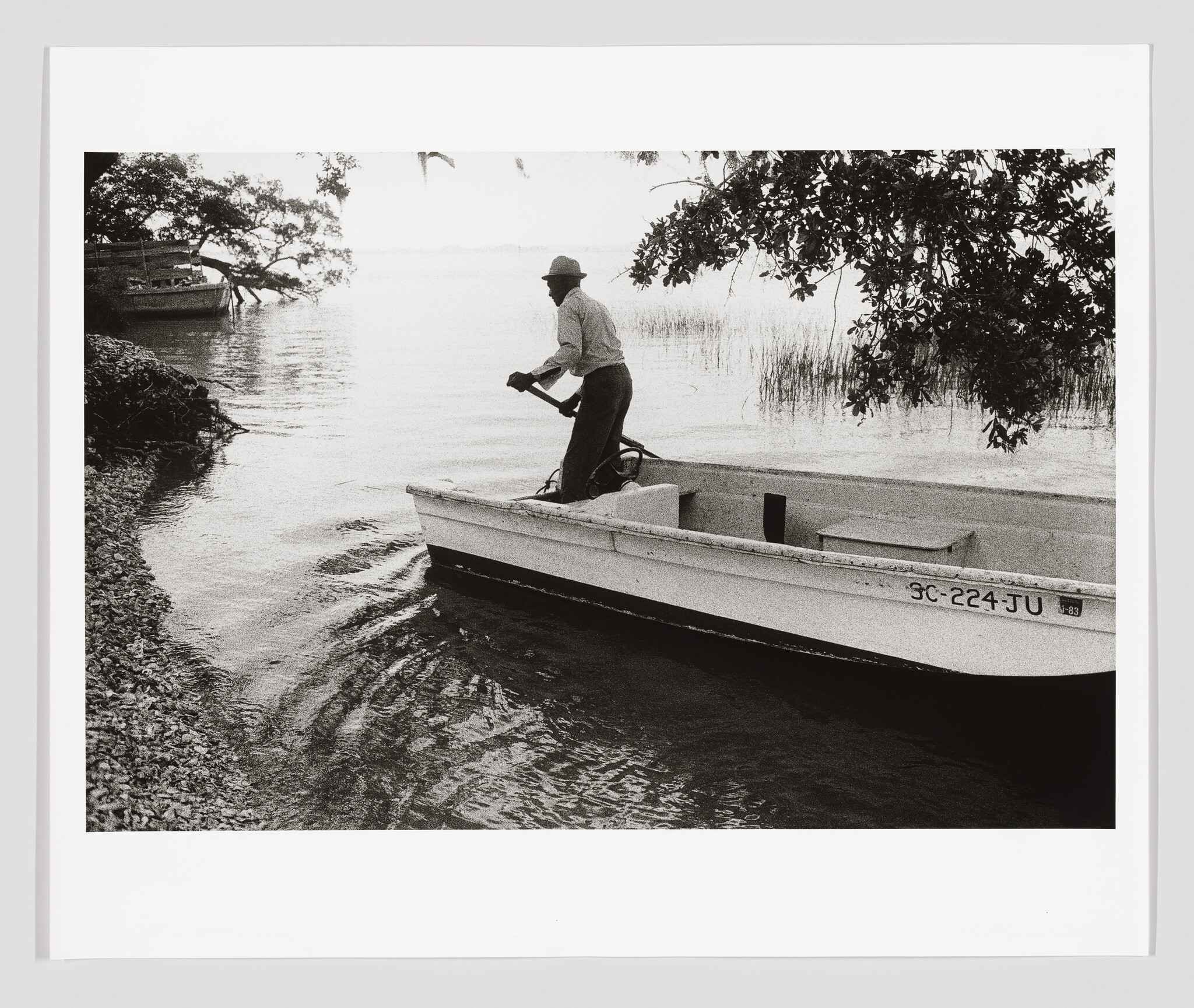 A man stands in a small boat pushing with an oar near a tree-lined shore.