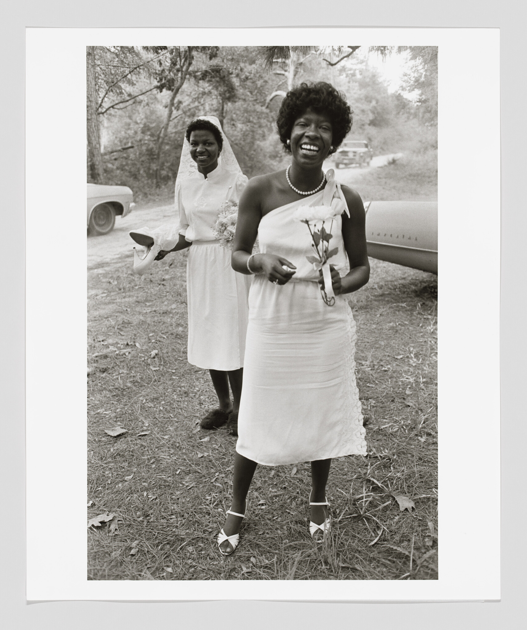 A smiling woman in a dress holds flowers while a veiled bride stands behind her holding shoes.