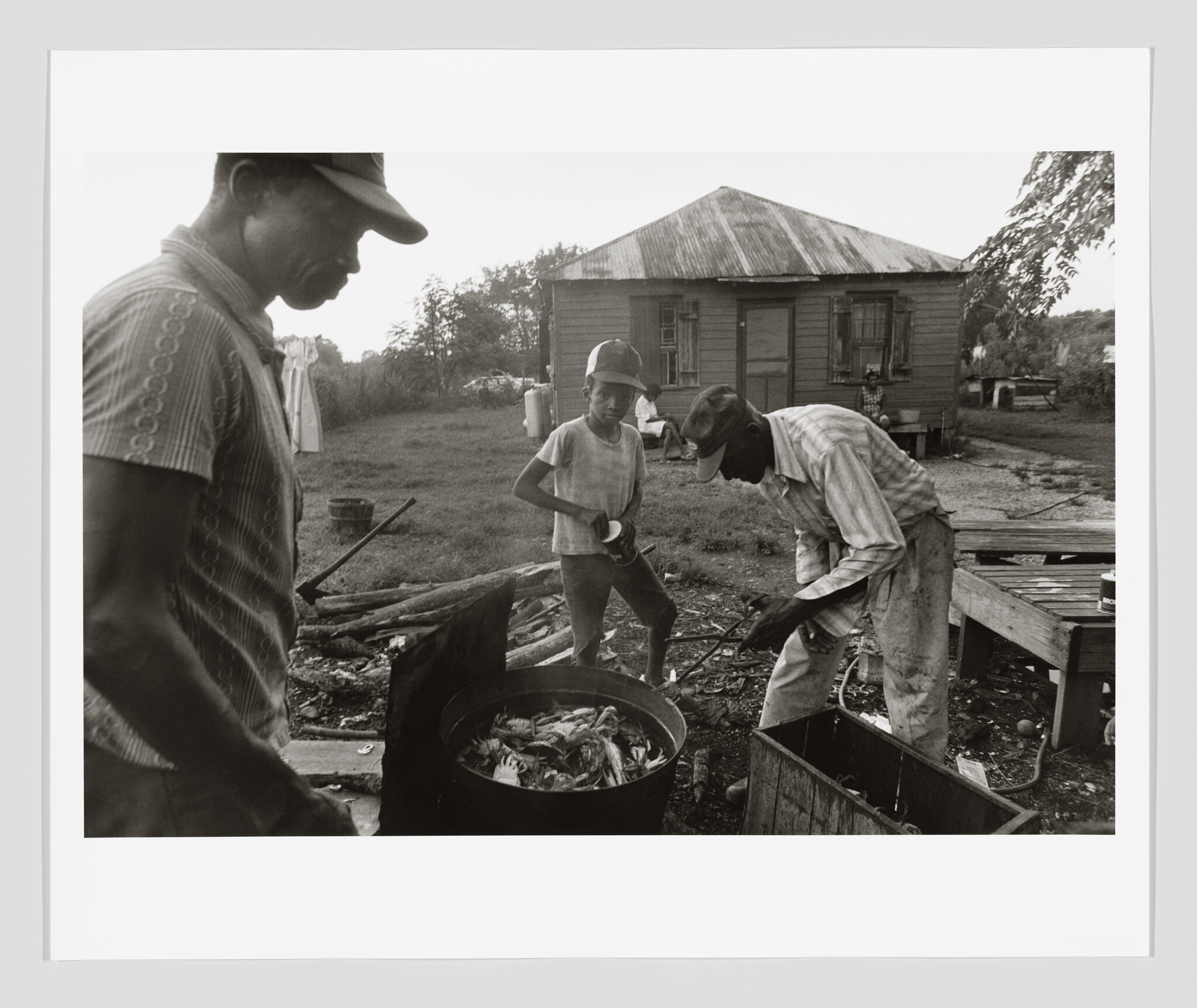 Three people stand beside a large pot outdoors as a man stirs seafood while a boy watches.