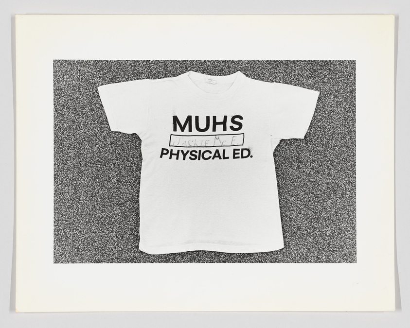 White T-shirt with "MUHS PHYSICAL ED." print, laid flat on a speckled background.