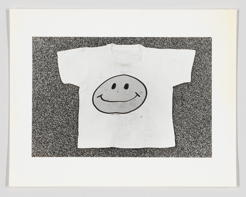 White t-shirt with a smiley face design on a speckled background, framed and displayed as art.