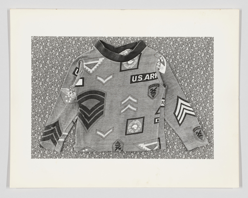 A child's grey camouflage shirt with various military-style patches on a patterned background.