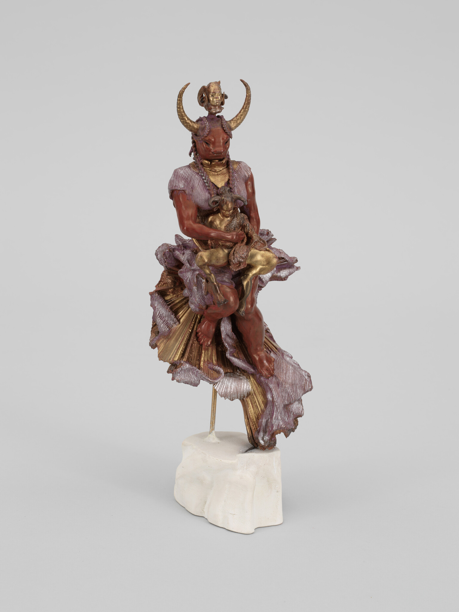 A horned humanoid sculpture wearing a flowing skirt holds a small gold animal while standing on a white base.