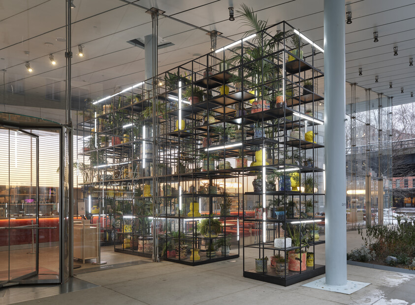 A tall metal shelving unit filled with potted plants and lit tubes stands inside a glass-walled lobby.