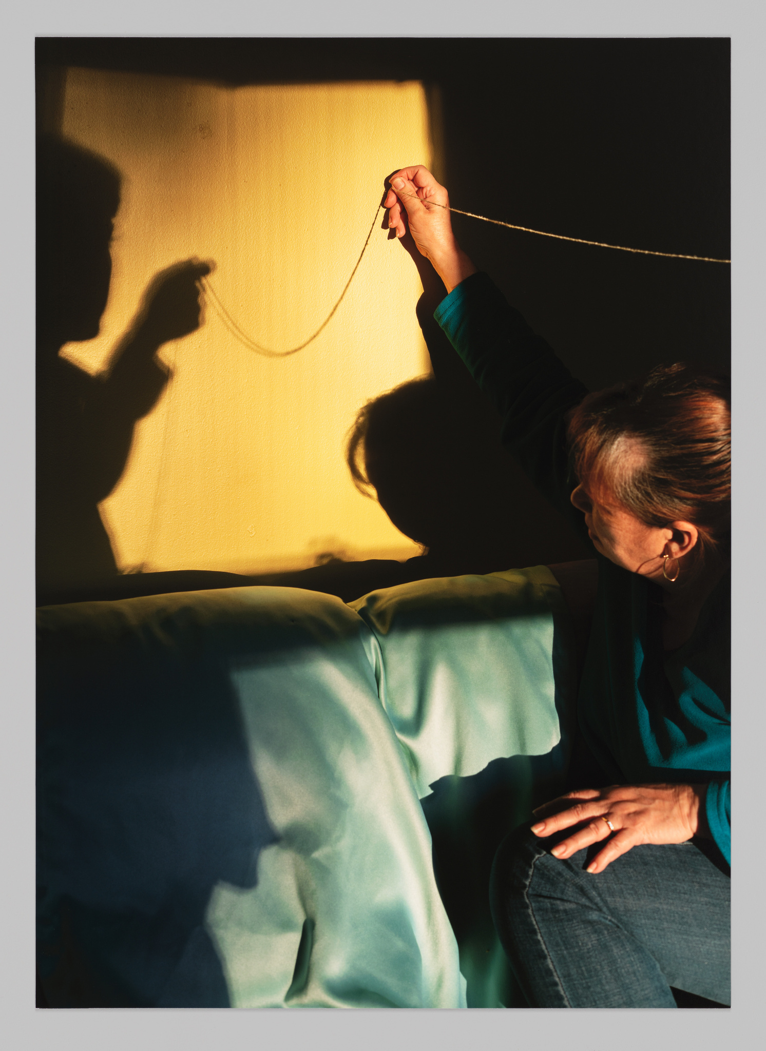 A string is held by two figures. One figure is only visible through their shadow, which darkens a square of light in the image background. The other figure is sitting on a blue couch with their arm stretched above them holding the string. The shadow of their face also appears in the square of light. 