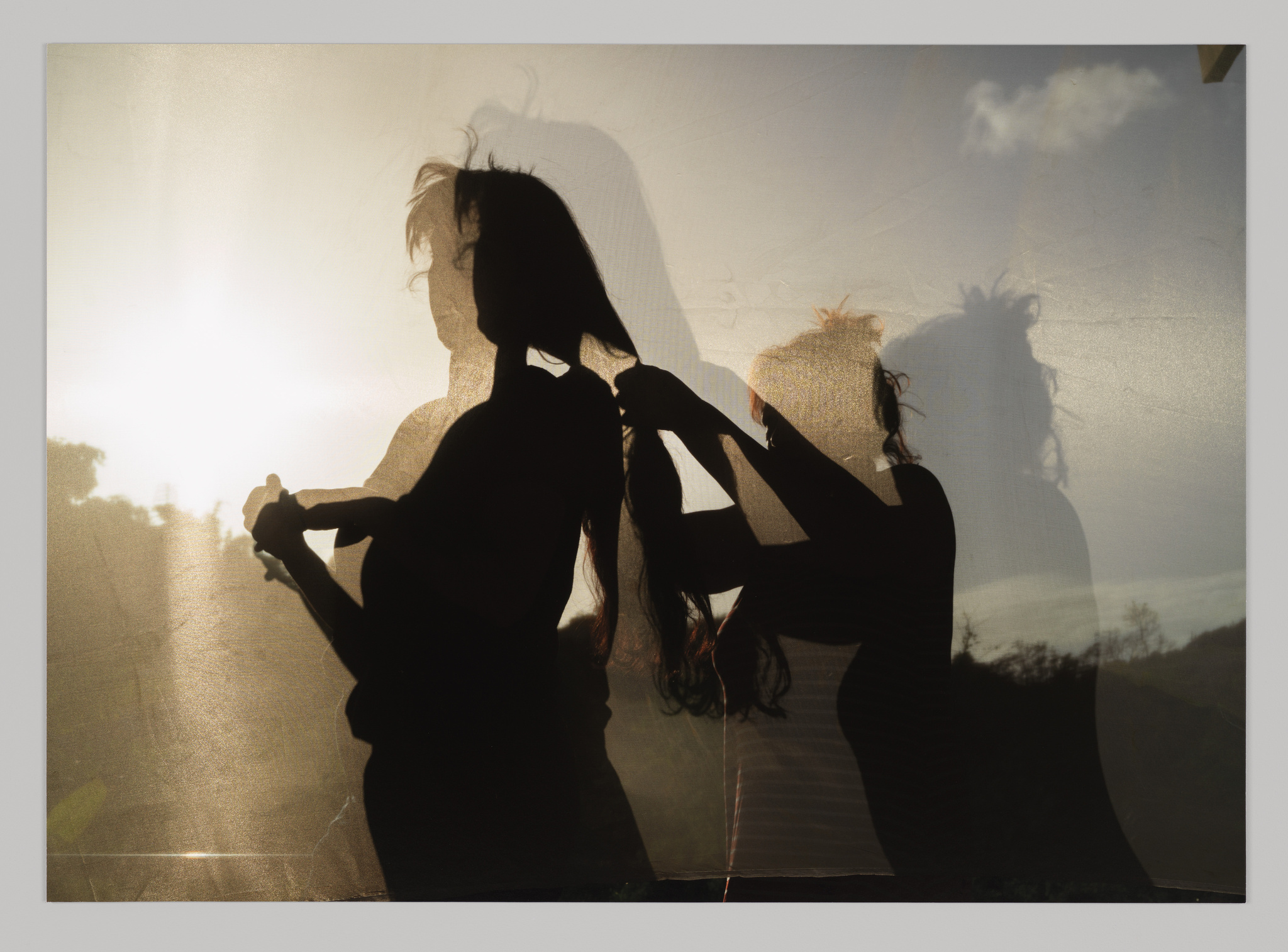 A photo of two figures in silhouette. The shorter figure is braiding the taller figure’s hair.