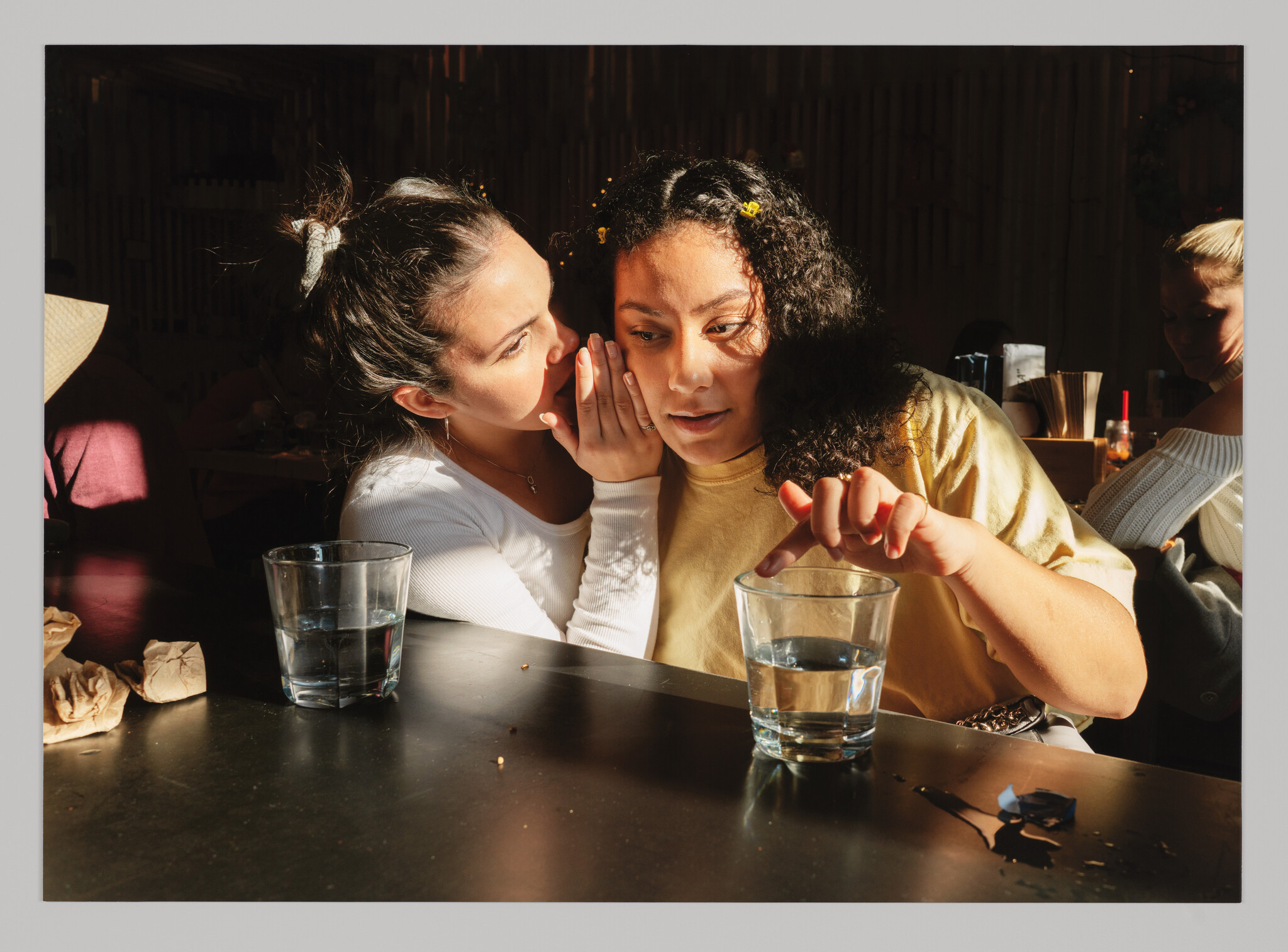 Two women sit at a bar with cups of water. One whispers into the other’s ear while she touches the rim of her glass.