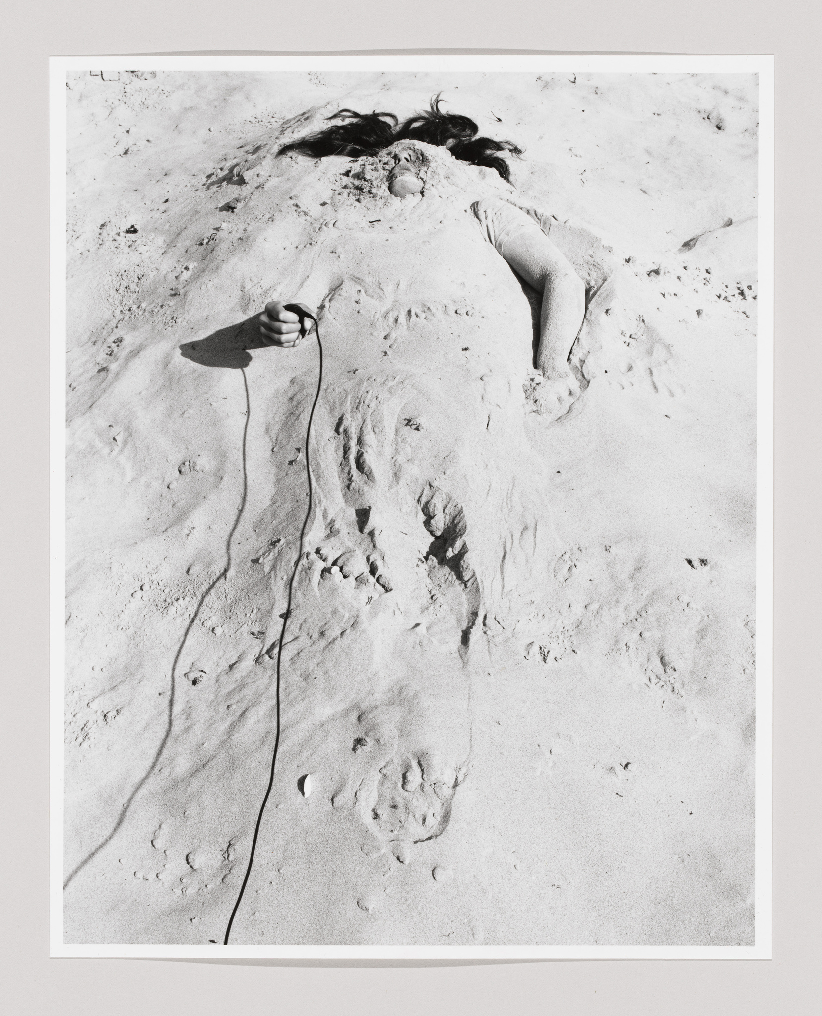 Person buried in sand with only their head and one arm visible, holding a cord. Black and white photograph.