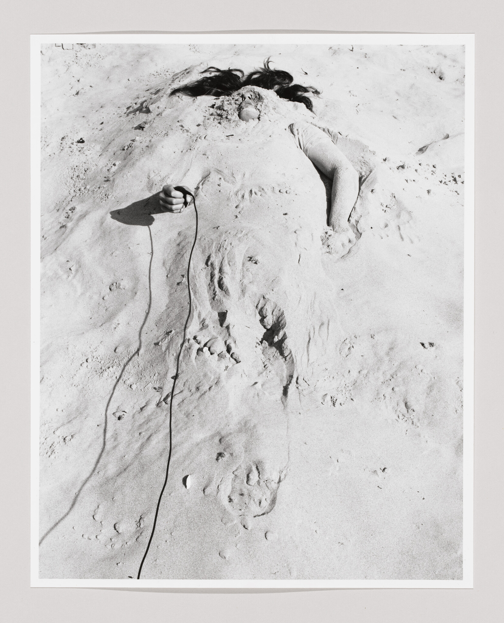 A person buried under sand with hair visible and one hand holding a cord.