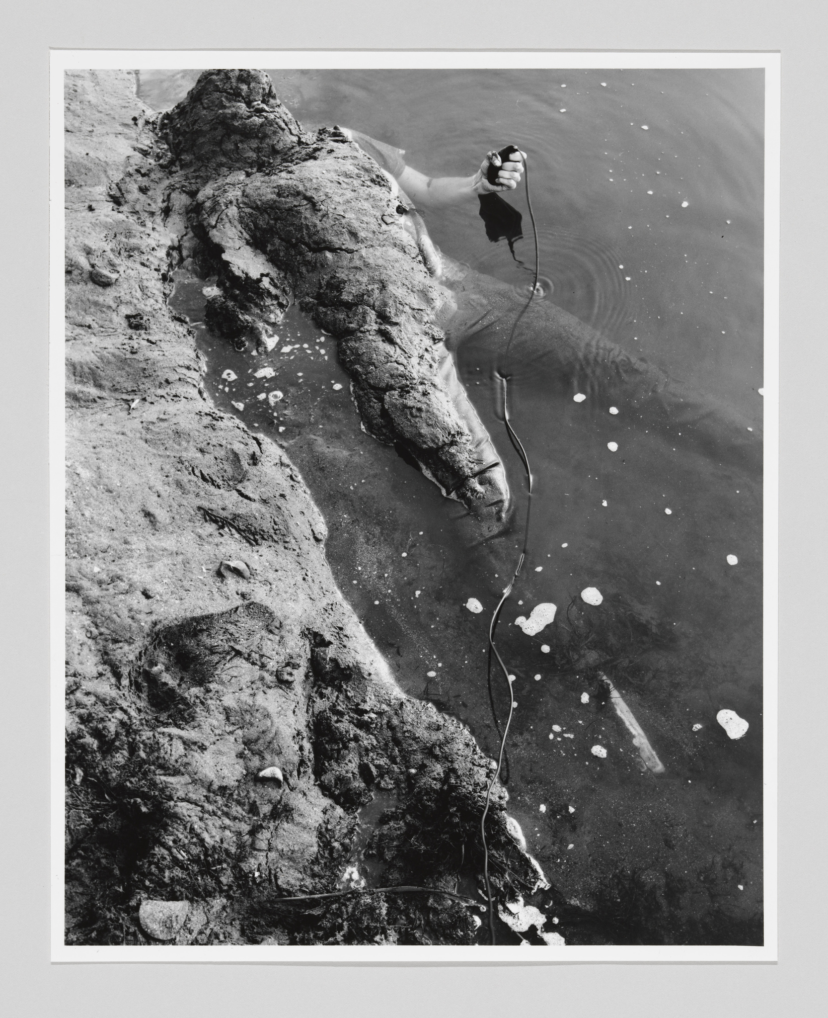 A black and white photo of a person half covered in dirt, half submerged in water, holding a cord.