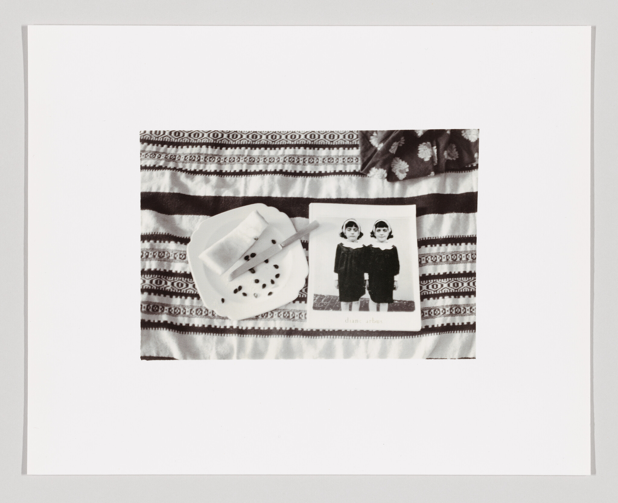 A black and white photo of twins lies on a patterned cloth beside a plate with a napkin.