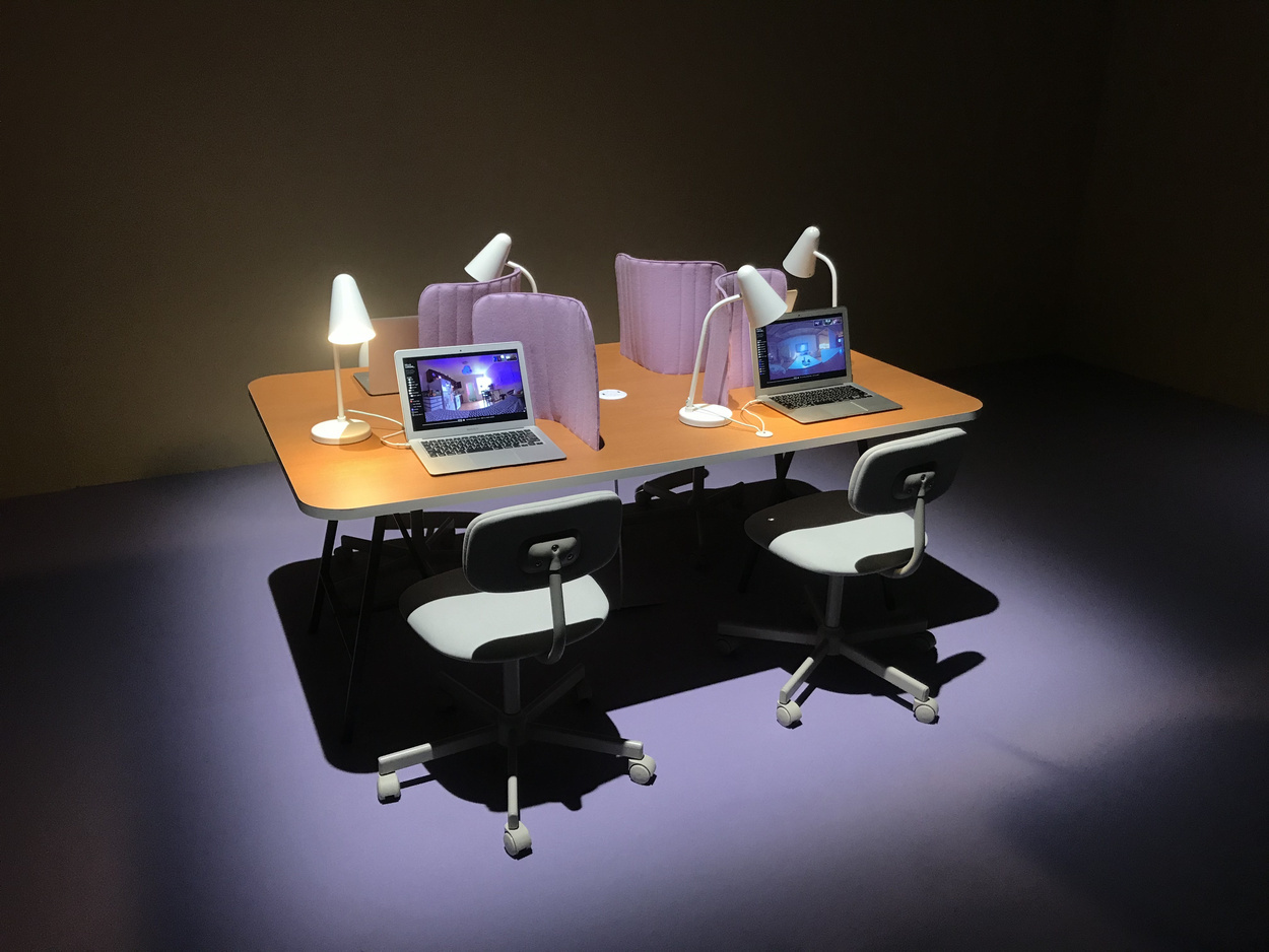 A modern workspace with two laptops open on a wooden table, flanked by white desk lamps and pink fabric dividers. Two ergonomic chairs with wheels are positioned at the table, all under a warm, focused lighting in a room with dark walls.