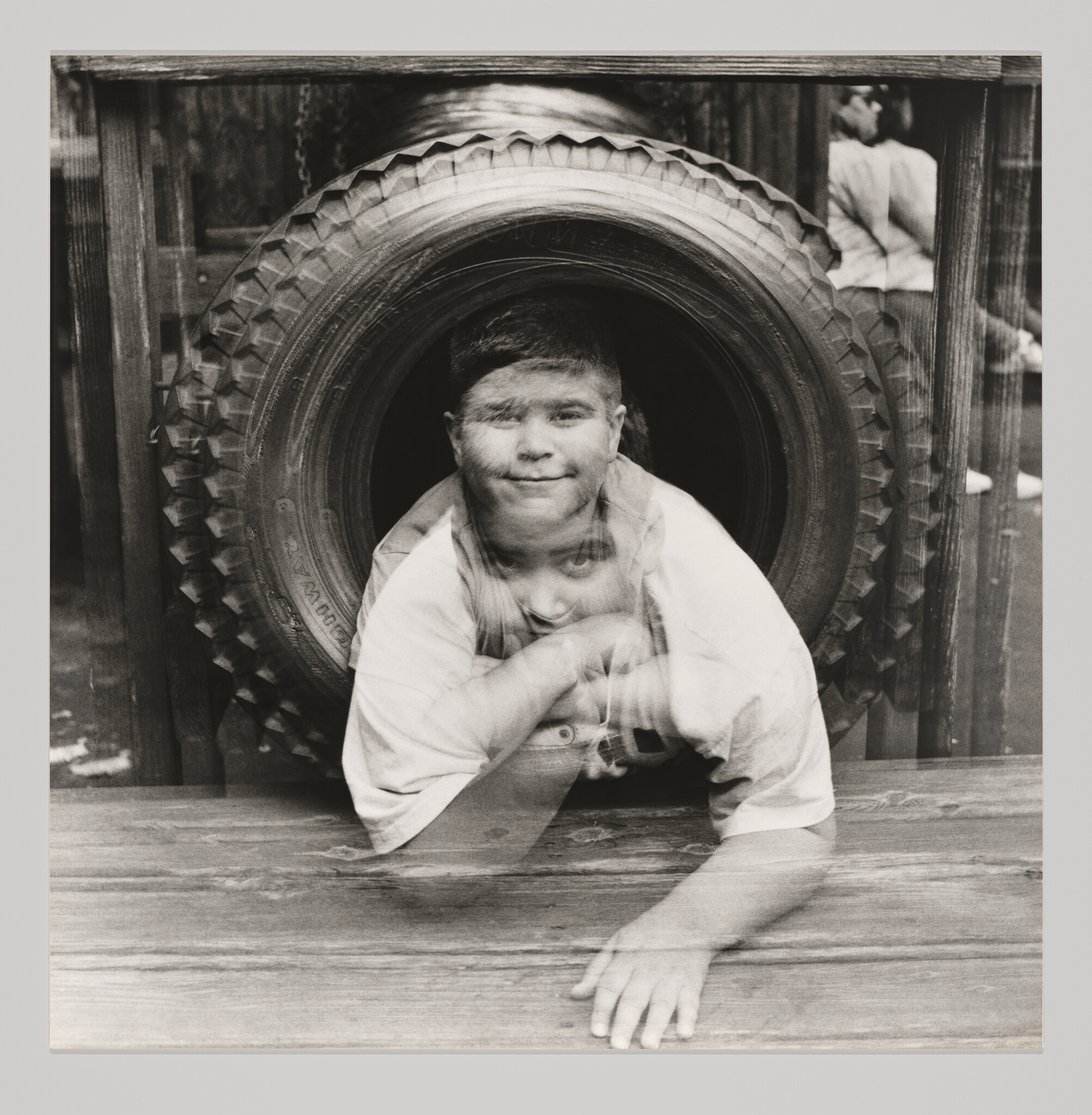 A double-exposed boy crawling through a large tire, smiling and resting an arm on wooden planks.