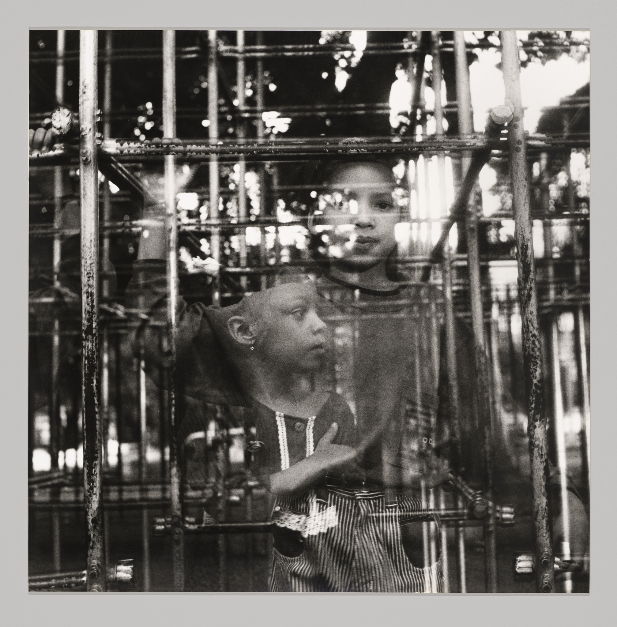 Two children are seen through a metal structure, creating a layered, reflective effect in this black and white photograph.