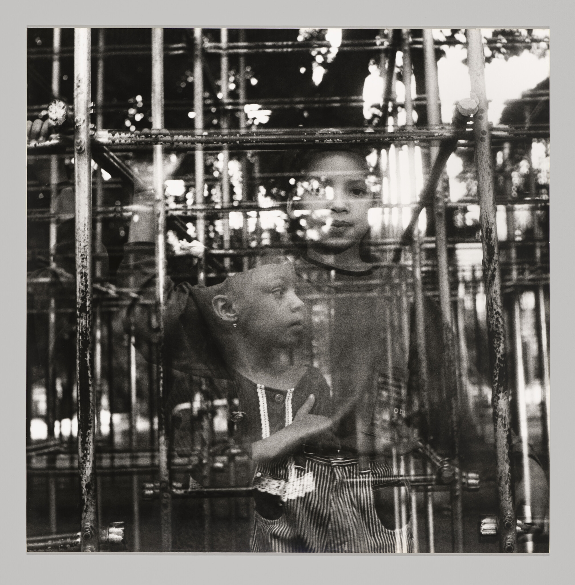 Two children stand behind metal bars and look outward through layered reflections.