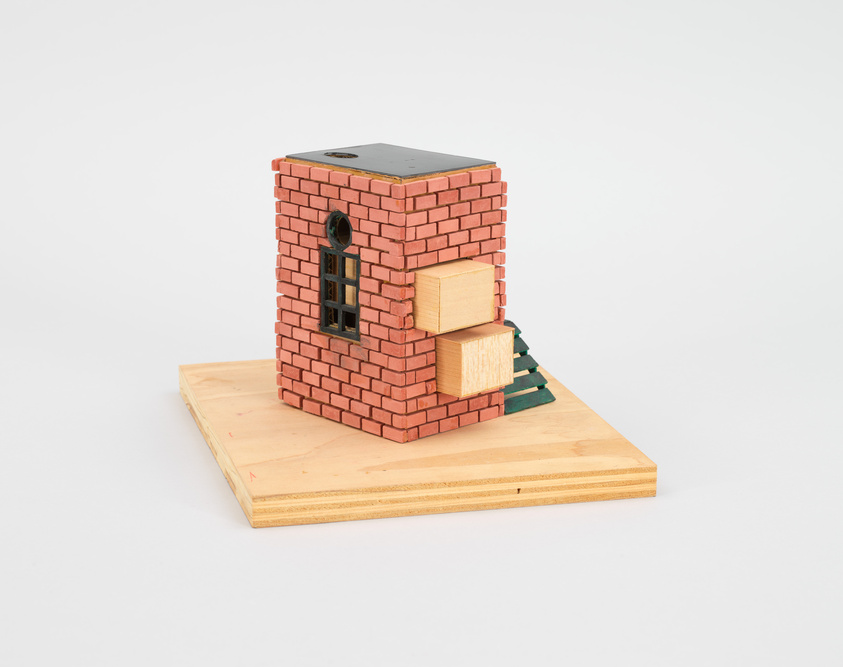 A small brick model building with a window and protruding wooden blocks, set on a wooden base.