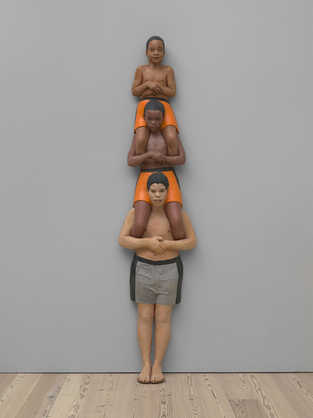 Three people stacked vertically against a wall, each sitting on the shoulders of the one below.
