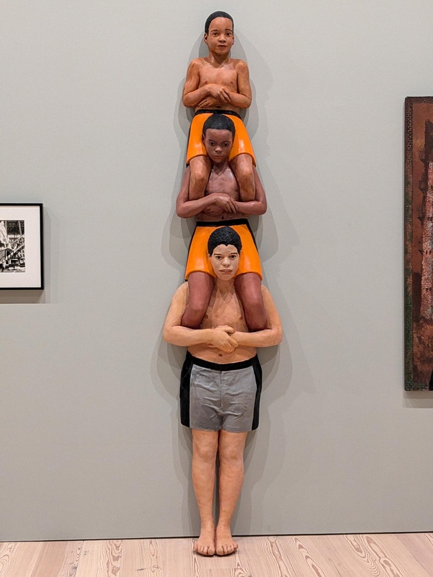 Three stacked sculptures of boys in orange shorts, each sitting on the shoulders of the one below, displayed against a gallery wall.