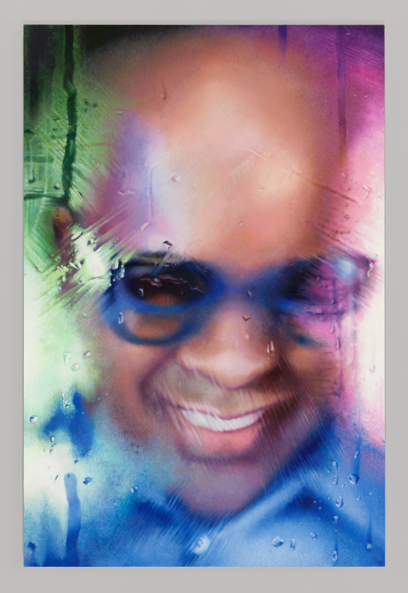 Smiling person with glasses behind a rain-streaked, colorful glass surface, creating a blurred, abstract effect.