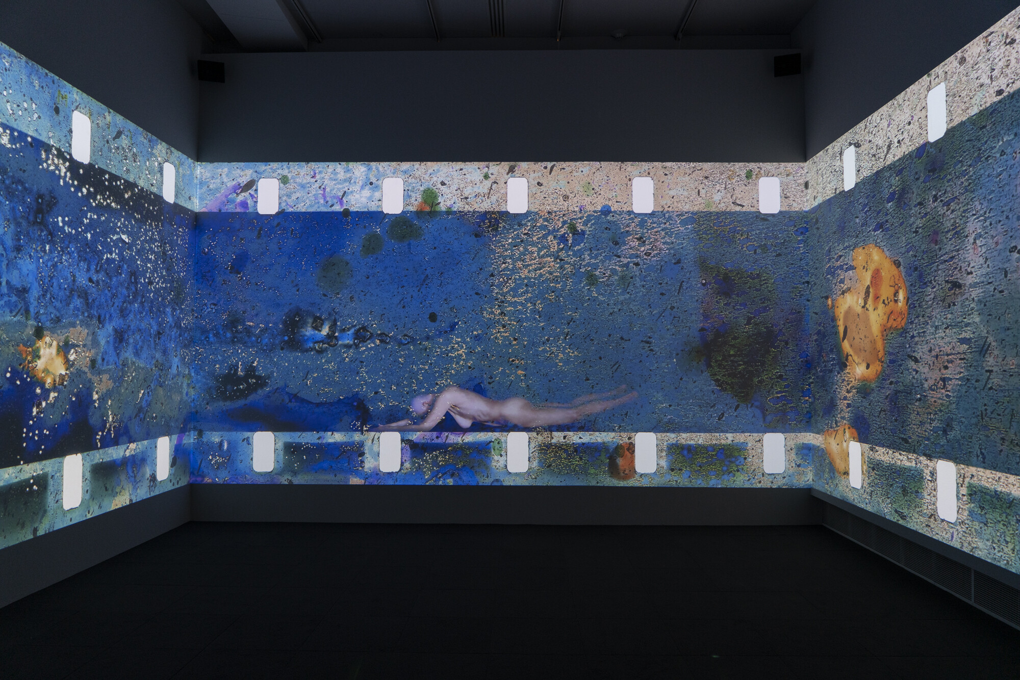 A large projection of a person swimming underwater is displayed on three walls in a dark room.