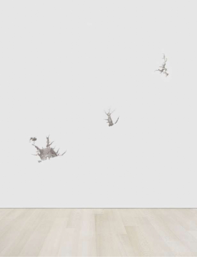 Three small, abstract figures appear to float on a white wall above a light wooden floor.