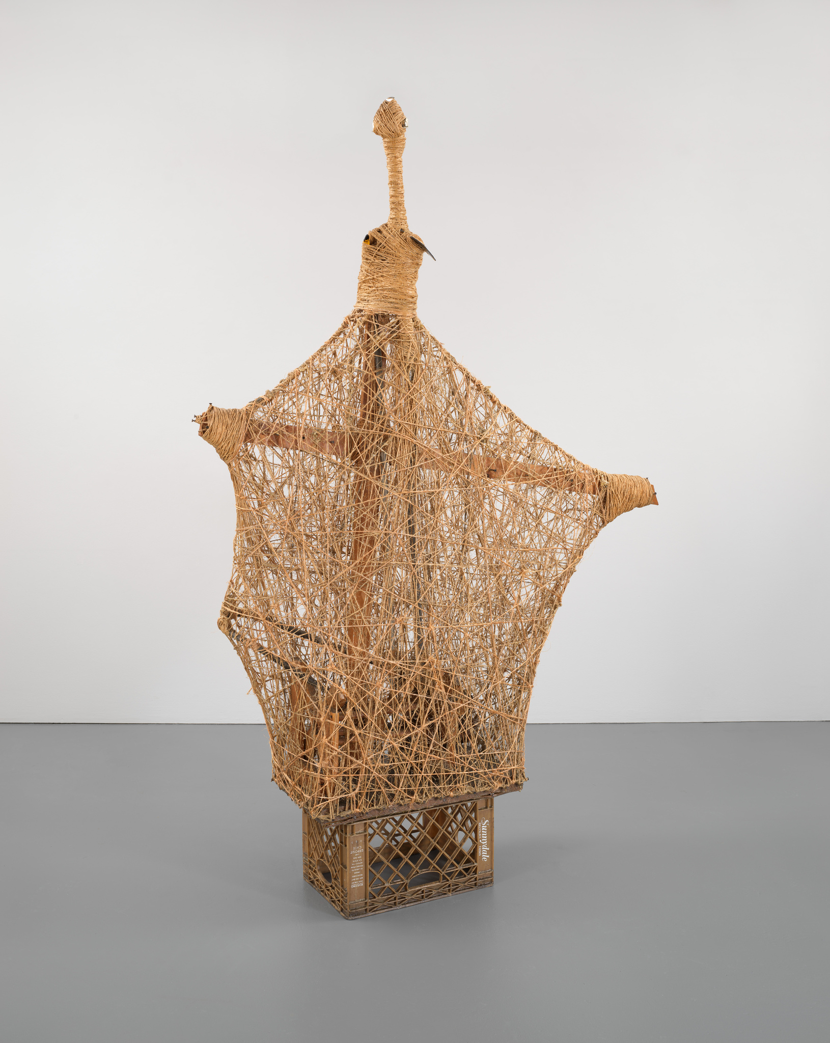 A tall, abstract sculpture made of intertwined twine, resembling a bird with outstretched wings, stands on a crate in a minimalist gallery.