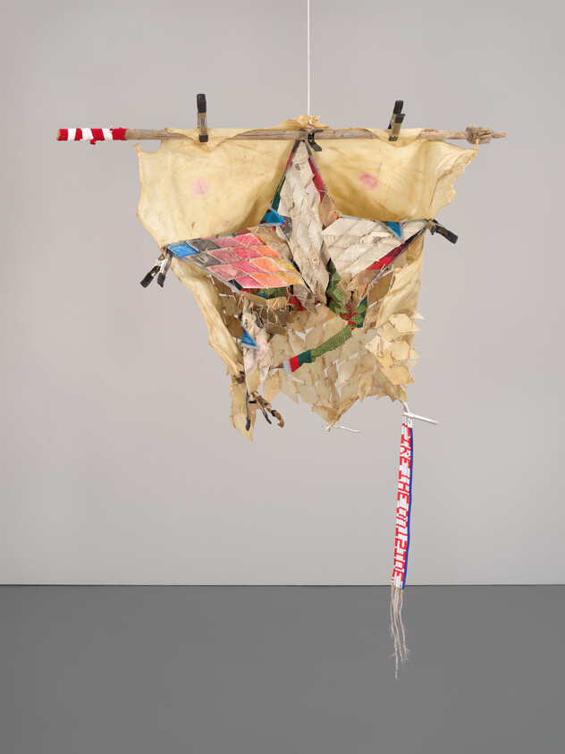 Abstract hanging sculpture with colorful geometric shapes and tattered fabric, displayed in a gallery setting.