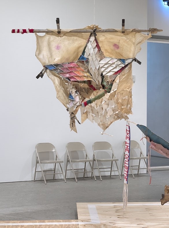 Abstract hanging sculpture with torn fabric and colorful geometric patterns, set against a gallery wall with folding chairs.