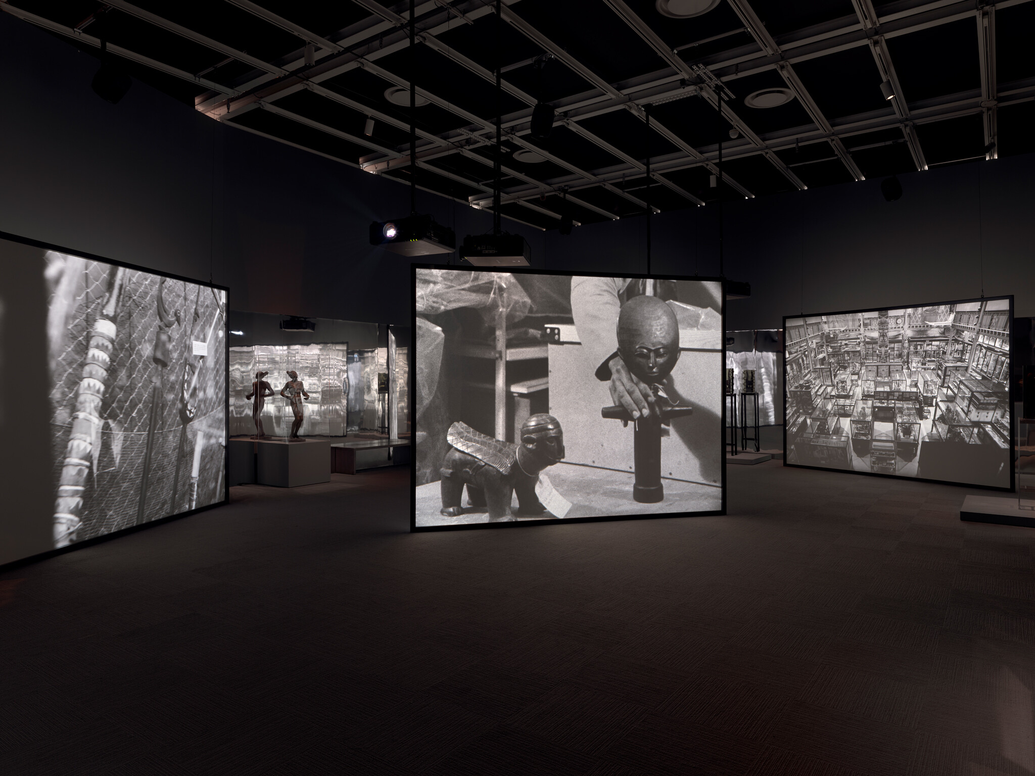 Large screens in a dark room display black and white images of museum artifacts and a person handling objects.