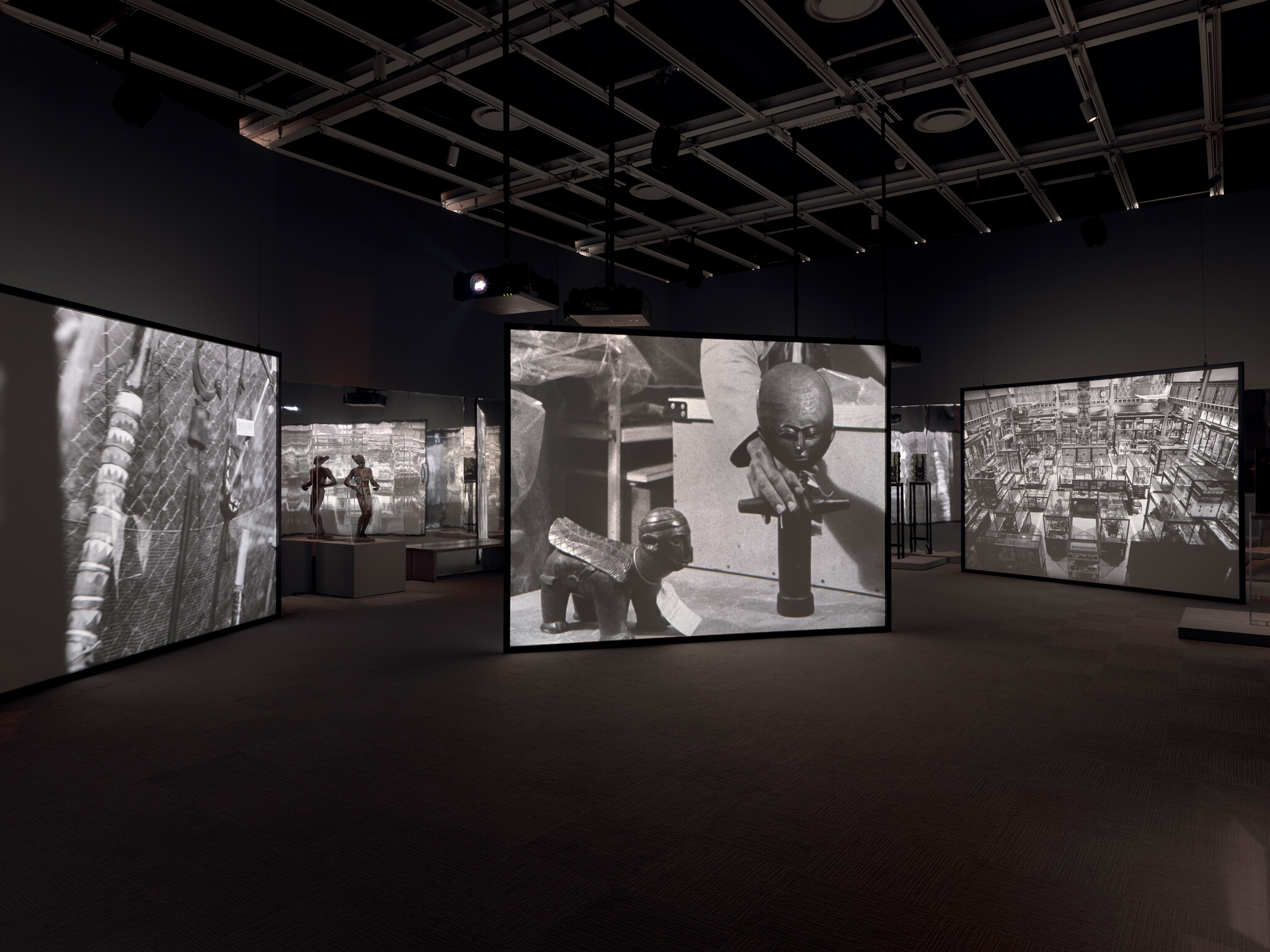 Large suspended screens in a dark gallery display black-and-white images of hands arranging small sculptures.