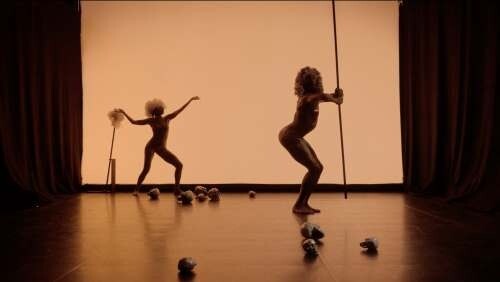Two silhouetted dancers perform on a dim stage, one holding a pole and the other balancing with a cane.