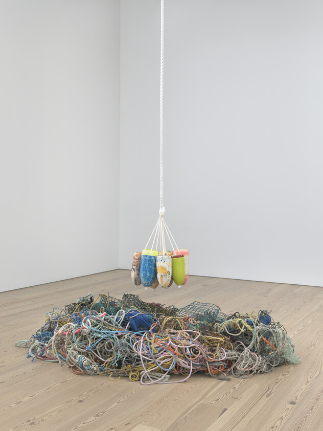 Colorful buoys hang above a pile of tangled ropes and nets on a wooden floor.