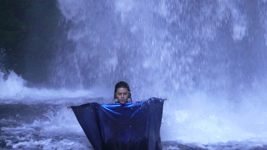 Person standing under a waterfall, wrapped in a dark blue fabric, with eyes closed and arms outstretched.