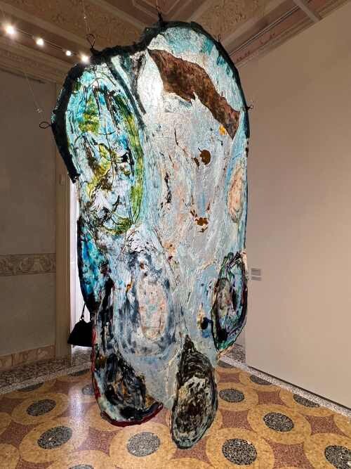 Large, abstract hanging artwork with swirling blue, green, and brown colors in a gallery setting.