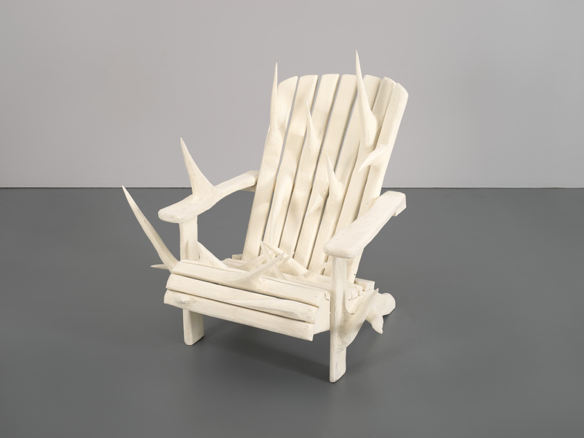 A white wooden chair with sharp, thorn-like protrusions on the seat, backrest, and armrests, placed on a gray floor against a plain background.