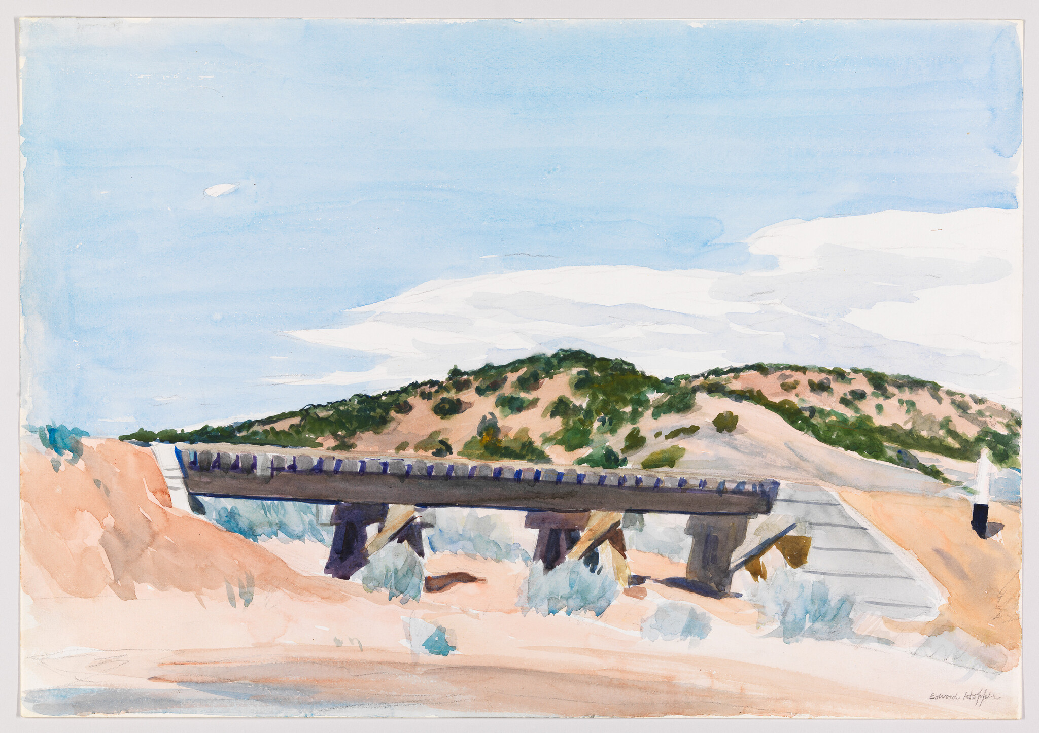 A wooden bridge crosses sandy dunes with green shrub-covered hills under a blue sky.