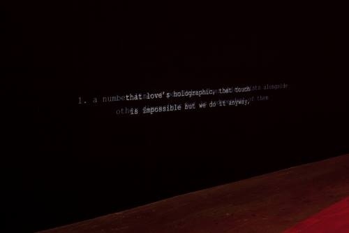 Text on a dark wall reads: "1. a number that moves holographically that touches the impossible but we do it anyway."