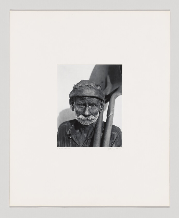 Older man with a worn hat and beard carrying a shovel over his shoulder.