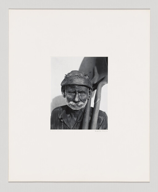 Older man with a worn hat and beard carrying a shovel over his shoulder.
