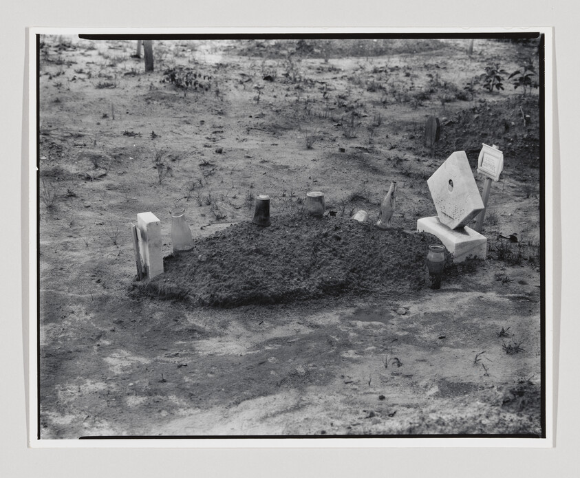 A freshly covered grave mound surrounded by small markers and jars in a bare sandy area.