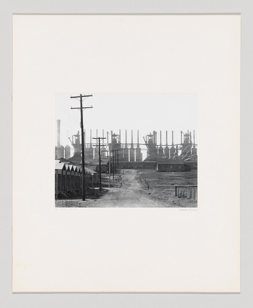 Dirt road leading past workers' housing toward large steel mill blast furnaces and smokestacks.