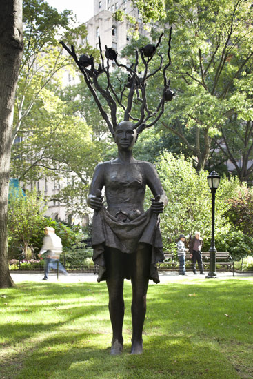 Bronze statue of a woman with tree branches and birds on her head, standing in a park with people walking in the background.