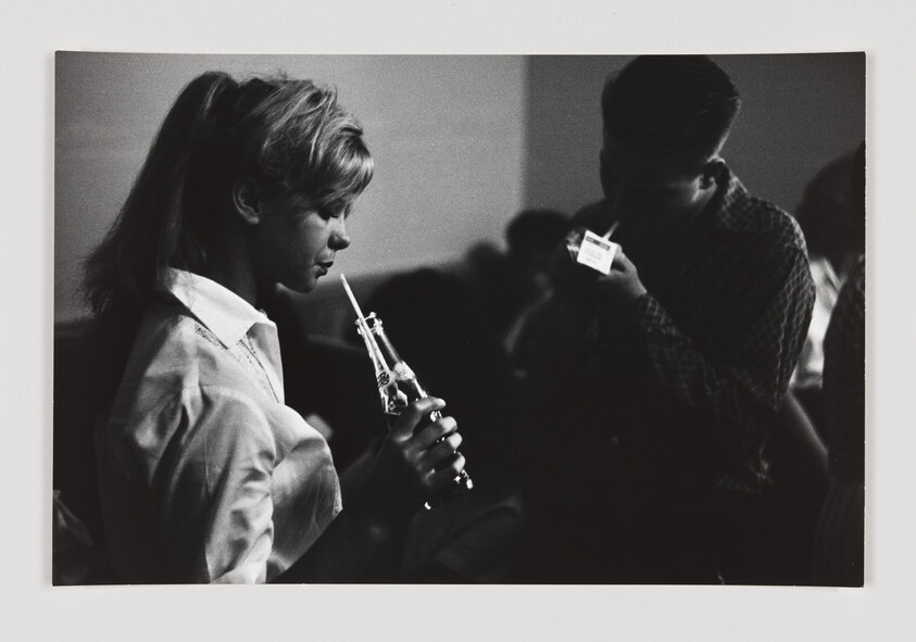 A young woman drinks from a glass bottle with a straw while a man lights a cigarette nearby.