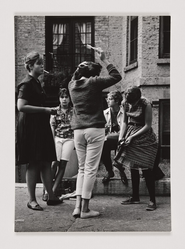 Five young women stand and sit outside a building, one fixing her hair with a brush, others watching.