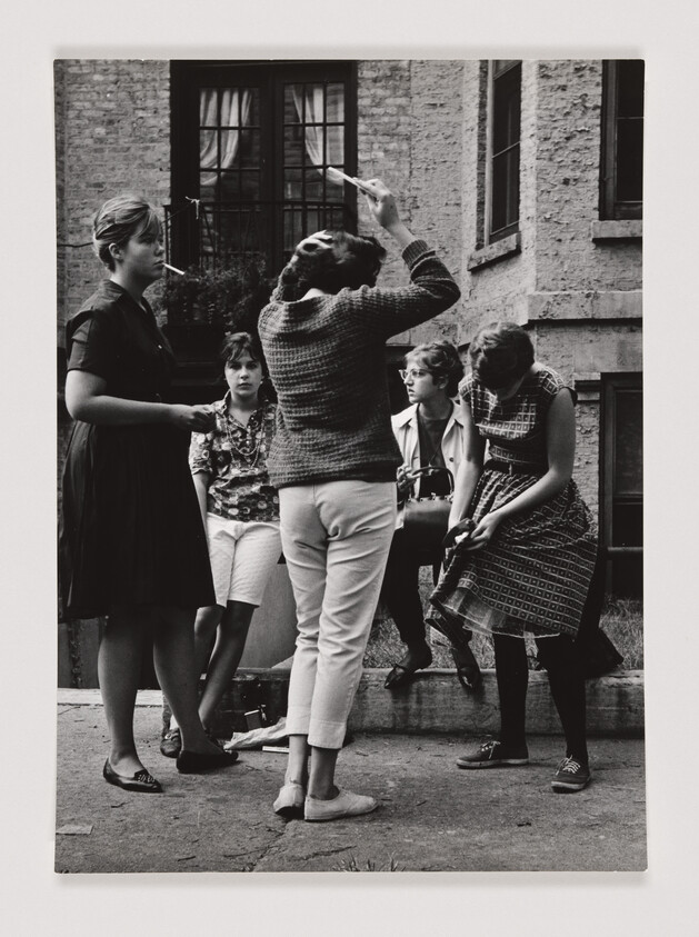 Five teenage girls gather outside a brick building as one raises a hand and others talk.