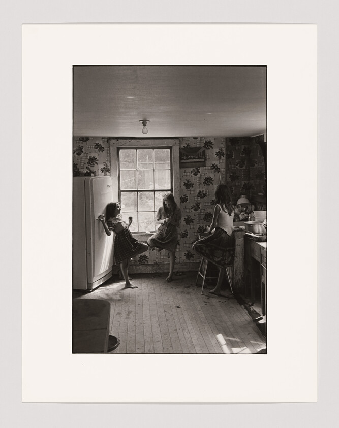 Three young girls in dresses talk and relax together in a small, old-fashioned kitchen near a window.